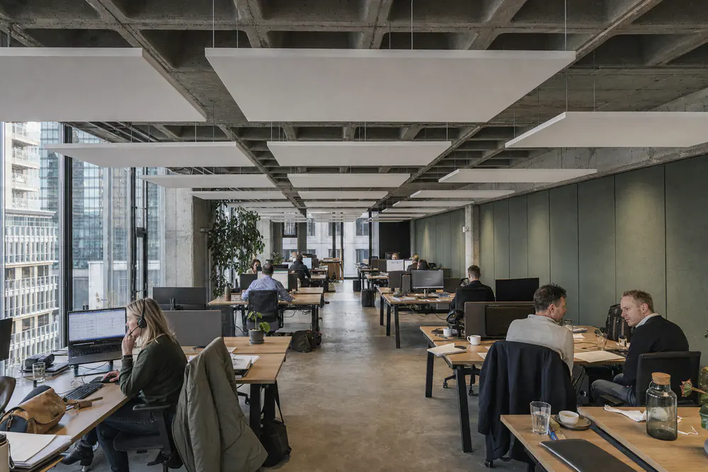 Open kantoor in een betonnen kantoorgebouw met witte plafondeilanden boven de werkplekken en een wand van groen vilt